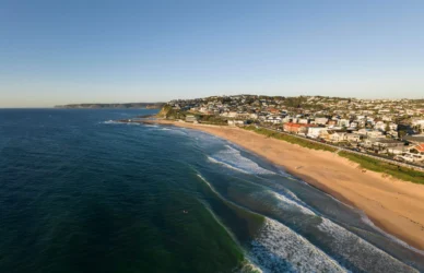 Coastal view from hotel room, featuring ocean waves, sandy beach, and nearby town under clear blue sky.
