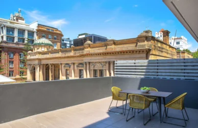 Enjoy the sun on a private hotel balcony with city views, featuring a dining table and two chairs, under a clear blue sky.