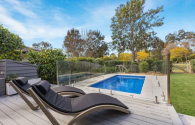 Private outdoor pool with glass safety fence, two black sunloungers on timber deck and leafy garden view
